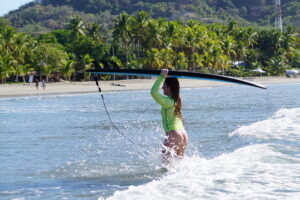 Costa Rica Surf camp surfer girl walks out of the water with her board in the tropical water in front of a palm tree lined beach at the Samara surf camp!