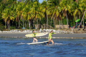Samara surf camp participants walk out of the water in front of a beautiful palm-lined shore