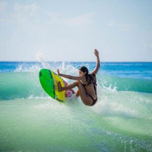 Alexxa Elseewi, surfing in her black bikini, hits the lip of the wave on her surfboard in clear turquoise tropical water