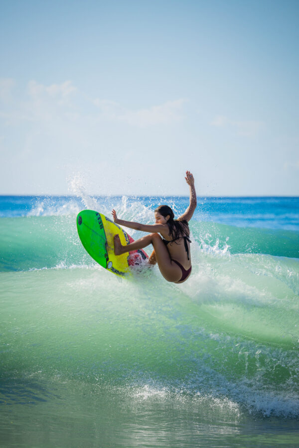 Alexxa Elseewi, surfing in her black bikini, hits the lip of the wave on her surfboard in clear turquoise tropical water